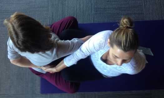 Two women perform a partner yoga pose, crossing their hands and holding each other’s arms.